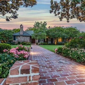 A brick walkway leading to a house with flowers in the foreground.