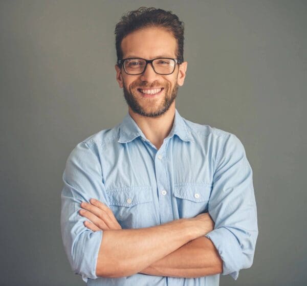 A man with glasses and beard standing in front of a gray wall.